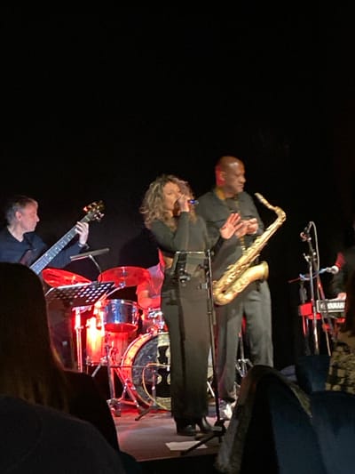 A musical performance is observed on a dimly lit stage in Cucuron, France. Five individuals are present on stage. A female vocalist with curly brown hair, wearing a dark top and skirt, stands center-left, holding a microphone to her mouth with her right hand while her left hand is raised. To her right, a bald male saxophonist, dressed in a dark shirt and trousers, holds a golden tenor saxophone with his left hand, his right hand extended forward. To the vocalist's left, a male bassist in a dark shirt plays a dark-colored bass guitar. Behind the vocalist, a drummer is partially visible behind a drum kit, which includes a bass drum with a dark, circular logo, all illuminated by prominent red stage lighting. On the far right of the stage, part of a "YAMAHA" brand keyboard and a musician's arm are visible. The foreground shows the backs of several audience members' heads, indicating a live performance setting. The background is uniformly black, directing focus to the performers on stage.