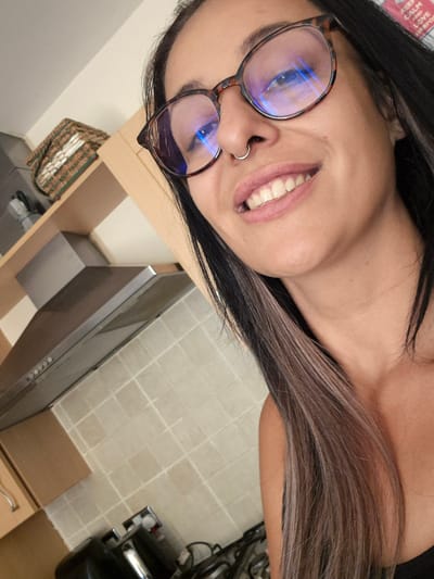 The image is a close-up, selfie-style photograph of a young woman with dark hair, some of which is highlighted with lighter streaks. She is wearing tortoiseshell-patterned glasses with blue light filtering lenses and a silver nose ring. She is smiling, revealing her teeth. The background is a kitchen, with tiled walls and a stainless steel extractor fan visible. A woven basket is on a shelf above the extractor fan. The image appears to have been taken indoors in a residential setting. The location is not specified within Bridlington, United Kingdom.