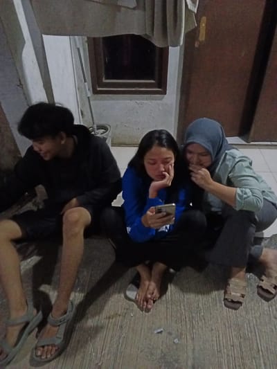 The image captures three young individuals sitting on a textured concrete floor, likely in an outdoor or semi-outdoor space in Bojong Gede, Indonesia. A young man on the left, wearing a black hoodie and shorts, is looking down and smiling. To his right, a young woman in a blue long-sleeved shirt and black pants is intently focused on her mobile phone. Beside her, another young woman, wearing a blue hijab and a striped shirt, is also looking at the phone and has her hand covering her mouth. The environment appears rustic, with a slightly weathered wall and a wooden door visible in the background. Shadows suggest the presence of artificial lighting. The ground is a light gray concrete with visible lines and some small debris. The individuals are casually dressed, and their postures indicate a relaxed interaction.