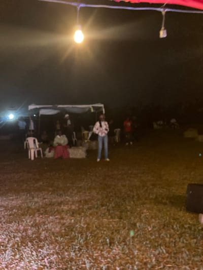An outdoor night scene in Murera ward, Kenya, depicts an assembly of individuals under artificial illumination. A single, brightly glowing incandescent lightbulb, suspended from an overhead structure, serves as a primary light source in the upper center, casting a visible starburst flare. Another strong light source emanates from the far left. The ground consists of dry, patchy grass. In the mid-ground, a white-canopied tent or gazebo shelters several people. One person is seated on a bale of hay or similar low structure, wearing a light green top and a red skirt. To their left, an empty white plastic chair and another occupied white plastic chair are visible. A central figure stands in the mid-ground, facing slightly right, dressed in a light-colored, patterned long-sleeved shirt and blue jeans. Additional indistinct figures are present under and around the tent, as well as scattered further in the dark background. A dark, rectangular object, possibly a speaker, is situated in the lower right foreground. The overall ambiance is dim, illuminated primarily by the visible light sources.