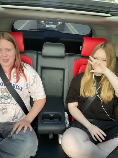 Two young females are seated in the back of a vehicle, likely an SUV, given the visible car seats and interior. The woman in the foreground is wearing a light pink pajama top with a teddy bear pattern and the word "OSHINE" is visible on it. She has light brown hair with pink streaks and is looking at the camera with her tongue sticking out. She is wearing a seatbelt.
The woman in the background is lying down and has long blonde hair. She is wearing a black t-shirt and a seatbelt. Her right hand is raised, and she is making a peace sign. She has a sparkly bracelet on her left wrist and is wearing earbuds. Her left leg is visible, and she appears to be wearing black denim shorts.
The interior of the car features red and black leather seats. A car's license plate reading "E745XY 18" is visible through the rear window of the vehicle the subjects are in. The weather appears to be sunny, and the time of day is likely daytime. The overall mood seems playful and casual.