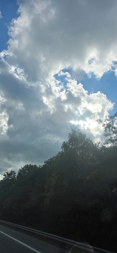 This media file captures a dynamic sky scene with a mix of bright sunlight and dark, heavy clouds. The sun is partially obscured by the clouds, creating a dramatic contrast. The sky transitions from a bright blue in some areas to a deep gray in others, indicating a changeable weather condition.
On the right side of the frame, a dense canopy of dark green trees borders the sky. The leaves are thick and form an almost solid wall of foliage, suggesting a forested area or a roadside lined with trees. The dark, almost black appearance of the trees might indicate they are in shadow, or the image was captured during a time of day when the sun was lower.
To the far right, a portion of what appears to be a road is visible. There are distinct white lines marking the lanes, suggesting it's a paved road. The road surface is dark, and there are some reflections on it, possibly from the sky or nearby light sources.
The overall impression is of a dramatic natural scene, possibly captured while traveling along a road through a wooded area. The time of day is likely either early morning or late afternoon, given the angle of the sunlight and the shadows cast by the trees. The weather appears to be mixed, with a potential for rain or just a dramatic, overcast sky. There are no people or specific objects clearly visible, focusing the attention on the interplay between the sky, the trees, and the hint of a road.