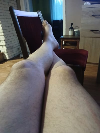The image shows a close-up view of a person's legs, crossed at the knee, resting on a maroon chair. The person is wearing no clothing below the knee. The background includes a brick wall to the left, a wooden dresser with drawers to the right, and a small wooden table with a white thermos and a plastic cup on it. A blue curtain is visible in the far background. The scene appears to be indoors, likely a residential setting. The location in the city of Gory, Russia, is not determinable from the visual information provided.