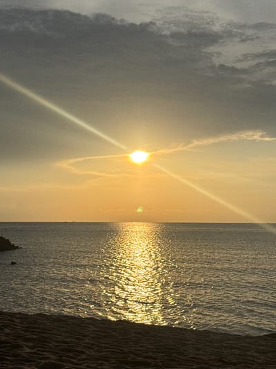 The image depicts a sunset view in Baan Wang Ta Kien, Thailand. The sun is low in the sky, casting a golden glare across the water. The sky is partially covered with clouds. The water surface is rippled, reflecting the sunlight. A shoreline or embankment is visible on the right side of the image.