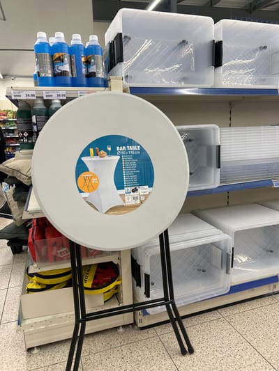 This media file captures an aisle within a retail store in Lampertheim, France, presented with a 90-degree clockwise rotation. The scene features various household and storage items.

In the prominent foreground is a light grey, round bar table top with a central label. The label reads "BAR TABLE" and specifies dimensions "Ø 80 x 110 cm," alongside a "MAX 75 kg" weight capacity. It also displays multiple translations like "BARTISCH," "BAROVÝ STOLEK," "TABLE DE BAR," and "TAVOLINO DA BAR." An illustration on the label shows the table with a white fitted cover, and a smaller image depicts its black metal base. The bar table's black, angular metal legs extend from beneath it, partially visible towards the right side of the frame.

Behind and to the left of the bar table, multiple clear plastic storage boxes with black clip handles are stacked on white shelves. These boxes feature a distinctive crisscross pattern on their interior bottom. Further left, on a lower shelf, are several blue bottles of liquid, possibly cleaning agents, with visible price tags of "39.98" and "39.95." Below the bar table, on a lower shelf, several red and yellow bags are stored, with one yellow bag clearly displaying the "DUNLOP" brand name.

The floor is a light-colored, checkered or tiled surface, consistent with a retail or warehouse setting. The overall impression is an organized display of products in an indoor shopping environment. No people are visible.