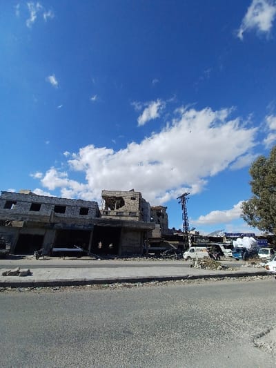 This image captures a street scene in Al-Qadam Municipality, Syria, under a clear blue sky with scattered white cumulus clouds. The foreground consists of an asphalt road and a damaged concrete sidewalk littered with debris.

To the left, a series of multi-story buildings appear severely damaged, characterized by exposed rebar, partially collapsed walls, missing roof sections, and empty window apertures. Their ground-floor entrances are dark and open. A single motorcycle is parked upright on the sidewalk directly in front of one of these compromised structures.

In the midground to the right, a tall metal utility pole stands. Further down the street, additional damaged structures are visible. Several vehicles are parked on the side of the road, including a white van, a blue truck, and a white sedan partially visible on the far right. Piles of rubble and debris are scattered along the roadside near these vehicles. A tree with green foliage is present on the far right edge of the frame. No individuals are clearly visible or engaged in specific actions within the scene.