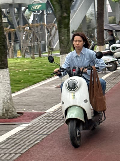 This image, rotated ninety degrees counter-clockwise, depicts an individual on a light-colored electric scooter in an outdoor urban or suburban setting during daylight. The main subject is an adult, likely with dark brown hair, wearing a light blue long-sleeved collared shirt and dark trousers. The person has earphones in and is looking towards the camera with a neutral expression. They are seated on a white or cream-colored electric scooter that features light green accents on its front panel. A brown bag is draped over the front of the scooter.

The scooter is situated on a paved surface. To its right, a reddish-brown path or road runs alongside a textured grey sidewalk, possibly tactile paving. To the left, a vibrant green grassy area is visible, interspersed with trees and some light-colored debris on the ground. In the background, beyond the grassy area, several buildings or structures can be vaguely discerned. Further back on the path, another person is partially visible next to a white scooter. The overall lighting suggests an overcast day or soft ambient light.

Two signs are visible in the background: a green sign on the left and a smaller yellow sign. Any text on them is too blurry and distant to be deciphered. The scene suggests a casual moment of travel or rest in a public outdoor space.