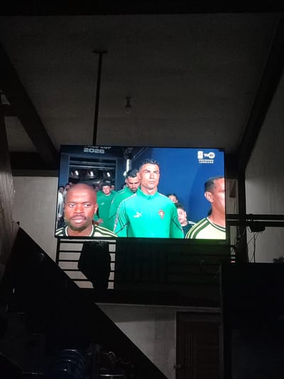 The image shows a large television screen displaying a soccer match or sports broadcast. The screen features the text "WORLD CUP 2026" and a logo for "TVRI LANGSUNG." Prominently featured on the screen are several soccer players, including Cristiano Ronaldo wearing a green jersey with the Portuguese crest. Other players are also visible in green jerseys. The scene appears to be indoors, with the television mounted on a stand. The background is dark, with some architectural elements visible, suggesting it might be a home setting in Cotseutui, Indonesia. The lighting is dim, with the television screen being the primary light source.