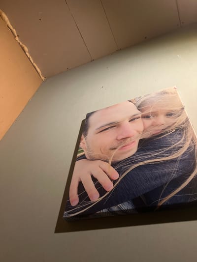 A framed photograph of two people is displayed on a pale green wall, positioned in the corner of a room. The photograph depicts a man and a child, with the child wrapped in the man's arms, their faces close together. The man has dark hair, a mustache, and is looking towards the camera. The child has blonde hair that is blowing across their faces. The photograph is partially obscured by a shadow cast on the wall to its right. The wall itself appears to be a light, muted green. In the upper left quadrant of the image, the junction of the wall and ceiling is visible. The ceiling is a light brown color, and a seam is apparent, with expanding foam insulation applied along the corner where the wall and ceiling meet. The lighting in the room is dim, with the photograph being the primary illuminated subject. The location in the city of Astoņi, Latvia is not discernible from the image content.