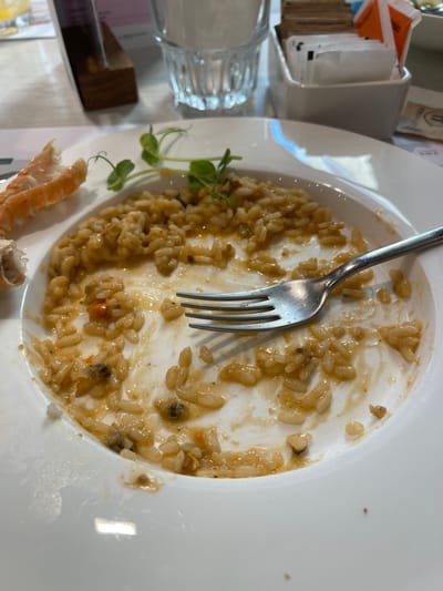 This media file shows a close-up, overhead view of a partially eaten plate of food, likely risotto, accompanied by a fork. The dish appears to be seafood-based, with visible pieces of shrimp or prawn and what could be mussels or other shellfish. The risotto itself is creamy and light brown in color, with visible grains of rice. A few sprigs of green garnish, possibly microgreens, are also present on the side of the plate.
The setting appears to be a restaurant, suggested by the presence of a water glass with condensation on the left side of the image and what looks like a restaurant menu or sugar packets in a holder in the background. The lighting is bright, indicating daytime. The overall impression is of a meal that has just been finished or is nearing completion. There are no people visible in the frame, only the aftermath of a meal.
The background is slightly out of focus, but hints at a clean, modern dining environment. The white plate is wide and shallow, typical of serving risotto. The fork is made of stainless steel and rests within the remaining food. The overall tone of the image is neutral, focusing on the food and its remnants. The location context of Noventa di Piave, Italy, suggests a European dining experience, possibly a trattoria or a more upscale restaurant serving Italian cuisine. There is no discernible text in the main portion of the image, though the out-of-focus background might contain some labels on the sugar packets or menu.
