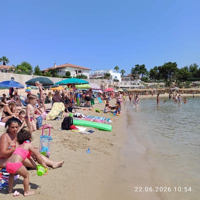 A wide shot captures a bustling beach scene under a clear blue sky. Numerous people, mostly in swimwear, are scattered across the sandy shore and wading in the shallow water. Beach umbrellas in various colors provide shade. Beach towels, inflatable mattresses, and children's toys are visible on the sand. Buildings with terracotta roofs and modern structures are situated in the background, bordering the beach. The timestamp "22.06.2026 10:54" is superimposed on the lower right corner of the image.