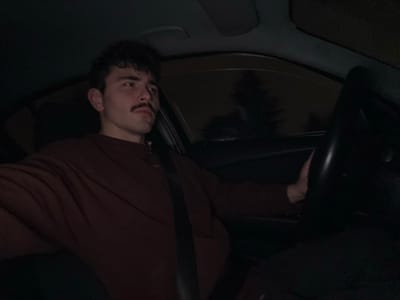 A person with dark hair and a mustache is inside a car, seemingly in the driver's seat, wearing a red-brown long-sleeved shirt and a seatbelt. The perspective is from an unusual angle, almost upside down, capturing the steering wheel and a partial view of the dashboard and front window with an indistinct reflection. The overall lighting is low, casting shadows within the car's interior.