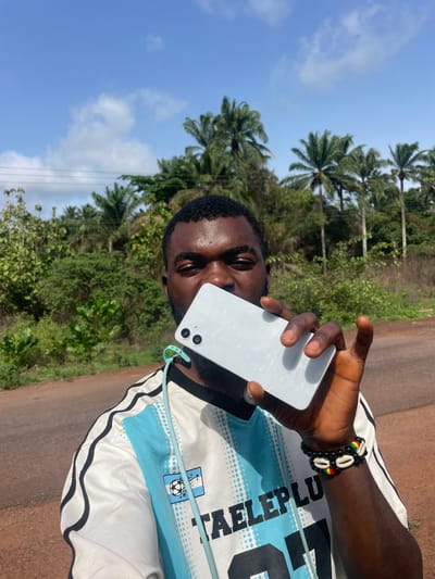 A man stands on an asphalt road in Ogbe, Nigeria, holding a light-colored smartphone to his ear. He is wearing a white and light blue jersey with black trim, adorned with a soccer emblem and the word "TALEPLU." A bracelet made of black beads, cowrie shells, and beads with red, yellow, and green stripes is visible on his wrist. The background features lush green vegetation with tall palm trees under a partly cloudy sky.