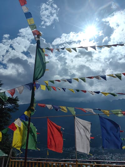 The photograph captures a view looking upwards at prayer flags strung across a bright, sunny sky with scattered clouds. The prayer flags are a mix of colors including red, yellow, blue, white, and green, and are triangular in shape, strung on lines supported by poles. The prayer flags are the main subject of the image. In the background, the sun is shining brightly, creating lens flares. Below the flags, a mountainous landscape is visible, with a town nestled in the valley. A wooden railing is present in the foreground, suggesting the photograph was taken from a balcony or viewing platform. The setting appears to be Siumsa, India, a location known for its Tibetan Buddhist culture and mountainous terrain. There are no people or animals visible in the image.