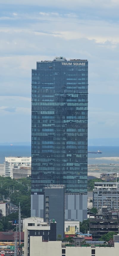 The image is a telephoto shot of the Trium Square building in Makati City, Philippines. The building is a modern skyscraper with a dark glass facade, featuring numerous windows that reflect the cloudy sky. The words "TRIUM SQUARE" are clearly visible in white lettering on the top of the building. In the background, the ocean can be seen with a ship sailing on it. Below the Trium Square building, other buildings of varying heights and architectural styles are visible, along with trees and some construction infrastructure. The scene appears to be an urban landscape under an overcast sky.