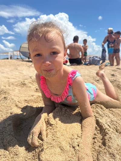 This media file shows a close-up, low-angle view of a young girl lying on a sandy beach, covered in sand. She is wearing a colorful swimsuit with a watermelon pattern and a pink ruffled trim. Her face is visible, with sand dusting her cheeks and mouth. Her eyes are looking towards the camera with a playful expression. In the background, partially blurred, are several adults and children also on the beach, some wading in the water, and others standing on the shore. The sky is blue with white, fluffy clouds, suggesting a sunny day. The scene appears to be a casual outing at the beach, likely in Blagoveschenskaya, Russia, given the location context. The time of day seems to be daytime, given the bright sunlight casting shadows.