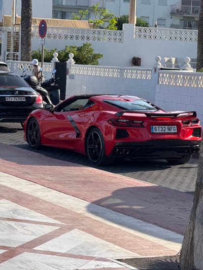 The image shows a red sports car parked on a street in Altea, Spain. The car has a license plate that reads "6132 NFG". Other vehicles, including a black Mercedes with license plate "T1-74798", are parked nearby. The scene appears to be outdoors, with sunlight casting shadows, suggesting it is daytime. The background includes white buildings with ornate detailing, palm trees, and lush greenery, indicative of a Mediterranean setting. The ground is paved with a patterned surface of pink, white, and grey stone. A blue and red traffic sign is visible, showing a red circle with a diagonal line over a blue square with a white arrow pointing left. The overall impression is a sunny day in a scenic Spanish town, with luxury vehicles on display.