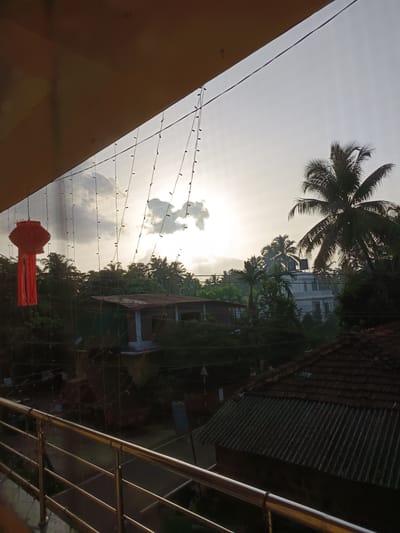 This is a landscape photograph taken from a balcony in Oxel, India, looking out at a cloudy sky with the sun attempting to break through, casting a diffused light. In the foreground, a silver railing lines the balcony. Hanging from strings are multiple small lights and a single red lantern, adding a festive touch.
Beyond the balcony, a dense collection of palm trees and other tropical foliage fills the middle ground, with several houses interspersed amongst them. One house, partially visible, has a brown tiled roof. Another, larger white building with a flat roof is also discernible. The overall impression is of a tropical or subtropical environment, possibly a residential area, with a hint of natural beauty and human habitation. The time of day appears to be late afternoon or early evening, given the quality of light.