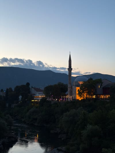 The image depicts a twilight scene in Mostar, Bosnia and Herzegovina, focusing on the Koski Mehmed Pasha Mosque and its surroundings. A tall, slender minaret pierces the sky on the left, adjacent to a dome-shaped structure illuminated by warm, orange light emanating from within the mosque. Below the mosque, a row of illuminated restaurants and cafes line the bank of the Neretva River, their lights reflecting on the water's surface. Dense foliage and trees dominate the foreground and mid-ground, with the dark silhouette of mountains forming the backdrop. The sky is a gradient of soft blues and grays, with scattered clouds near the horizon. No people are clearly identifiable.