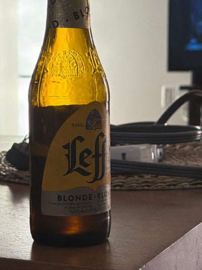 A single Leffe Blonde beer bottle, made of amber-tinted glass, stands upright on a dark, wood-grained table surface. The bottle's neck bears an embossed crest or emblem. Its primary label is yellow, featuring "Leffe" in a distinct gothic script, with "BLONDE" and "6.6% ALC./VOL." below, along with descriptive text in French about vanilla and clove notes. An illustration of a building, possibly an abbey, is visible on the label above the "Leffe" text, accompanied by "ANNO." The bottle shows condensation, indicating it is cold. It rests on a circular, woven placemat with a visible texture. The background is blurred, showing a bright, featureless wall or window on the left. To the right, a dark, rectangular electronic display (likely a television) with a faint blue reflection is visible. Behind the bottle, black coiled cables and a white power strip are indistinctly visible on the table. No people are present. The location in the city of Buford, United States, cannot be determined from the visual information provided.
