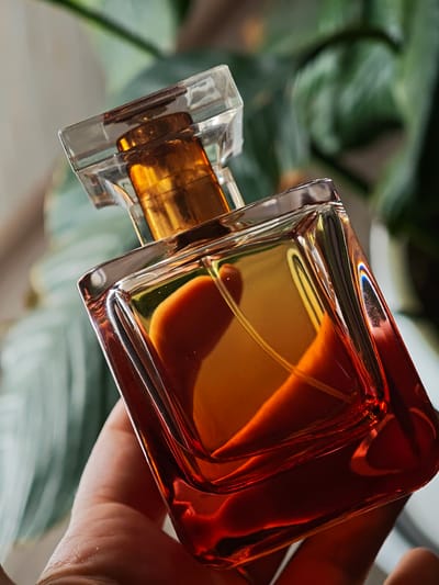 A hand holds a glass perfume bottle filled with amber liquid. The bottle has a clear, faceted cap and a golden atomizer. The background is blurred, showing green foliage and hints of a light-colored surface. The image is taken from a close-up perspective, emphasizing the texture and reflections of the perfume bottle. No location in Kursk, Russia is visible in the image.