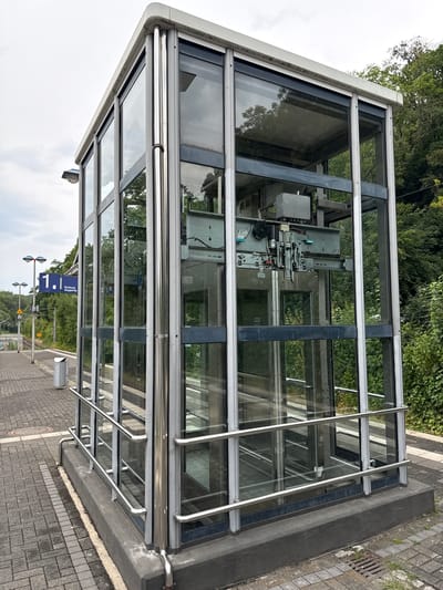 This media file shows an exterior view of a modern glass and metal elevator enclosure, likely at a train station or public transportation hub. The enclosure has a rectangular footprint with a sloped roof, constructed with a metal frame supporting large glass panels. Inside the enclosure, the mechanical components of an elevator are visible, including cables, pulleys, and motor housings. The elevator appears to be a service elevator, judging by the exposed machinery and lack of a passenger car interior visible.

The surrounding environment includes lush green trees and foliage in the background, suggesting a natural or park-like setting. The foreground is a paved area, possibly a sidewalk or platform, made of interlocking grey bricks. A metal railing runs along the exterior of the glass enclosure, providing a safety barrier. A blue and white directional sign with text is visible, reading "Richtung Wuppertal" which translates to "Direction Wuppertal" in German, indicating the location is in Germany and the elevator provides access towards Wuppertal. A small, yellow warning sign with a black symbol is also visible on a pole in the distance.

The lighting suggests it is daytime, with an overcast or cloudy sky visible in the upper portion of the image. The overall impression is one of functionality and modern infrastructure within a potentially scenic or well-maintained public space. There are no people visible in the image. The scene appears to be static, with no specific activity or event taking place other than the presence of the elevator structure itself.