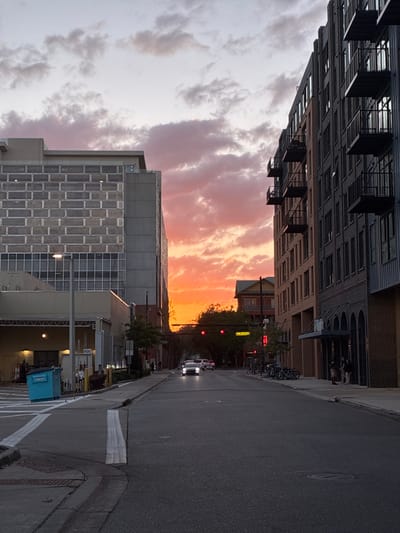 The image depicts an urban street scene in downtown Gainesville, Florida, United States of America, observed at dusk during a sunset, looking west.

The sky above is characterized by scattered clouds, transitioning from pale grey and white in the upper regions to prominent pink and orange hues at the western horizon, intensely illuminated by the setting sun.

On the left side of the street, a multi-story modern building dominates, featuring an upper facade composed of a grey, textured, rectangular grid pattern. Below this, large windows are visible. The lower section includes a covered vehicle entrance or exit bay, with a yellow "FDC" sign prominently displayed on a stand. A bright blue waste dumpster is positioned on the sidewalk near this bay.

On the right side of the street, a multi-story contemporary building with a dark grey exterior and numerous projecting balconies with dark metal railings extends into the distance. The ground floor features recessed arched openings and a section of lighter brown material. Several bicycles are parked in a cluster on the sidewalk in front of this building. Four individuals are standing on the sidewalk near the building's entrance, appearing to be in interaction.

The asphalt street, marked with white painted lines in the foreground, extends into the background. Two vehicles are visible in the distance, heading towards the viewer. The lead vehicle, dark in color, has its headlights illuminated. Further down the street, an intersection is visible, equipped with traffic lights, the primary signal facing the viewer displaying red. Silhouetted trees and the upper portion of a smaller, older-style building with a peaked roof are discernible beyond the intersection against the bright sunset.