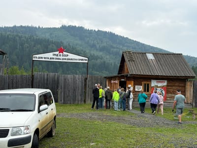 The image depicts an outdoor scene in Ust'-Kabyrza, Russia, featuring a wooden building with a rustic aesthetic and a group of people gathered outside. In the foreground, a white van, partially covered in mud, is parked on a gravel and grass area. A wooden fence runs along the left side of the scene, separating the area from a backdrop of a dense, forested mountain under an overcast sky.

A prominent sign with a red star is visible above the fence, displaying Cyrillic text that translates to "Labor in the USSR - is a matter of honor, a matter of glory, a matter of courage." To the right of the fence, the wooden building has a weathered appearance with a shingled roof. A banner hangs on the side of the building, featuring an image and Cyrillic text reading "We Remember! 1941-1945." Another sign with Cyrillic text is affixed to the wall near the entrance.

A group of approximately ten individuals, dressed in casual clothing, are positioned in front of and near the entrance of the building. Some are facing the building, while others are engaged in conversation. The overall atmosphere appears to be one of a gathering or a visit to a historical site. The environment is rural, characterized by the natural landscape and simple structures.