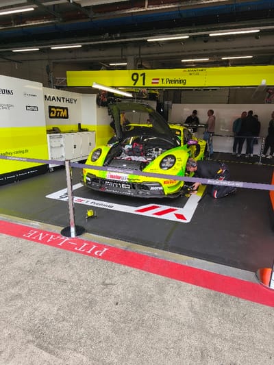 The image displays a pit garage at the Red Bull Ring circuit near Flatschach, Austria. A neon yellow-green Porsche 911 GT3 R race car, identified by the number 91 and "T. Preining" branding, occupies the center of the garage floor, which is covered with black modular matting featuring white and red markings. The car's front hood is open, revealing internal engine and chassis components. A single mechanic, dressed in dark racing overalls, kneels directly in front of the car's right side, performing maintenance or adjustments. A purple rope barrier, supported by chrome stanchions, delineates the immediate work area. In the background, on the right, approximately five to six casually dressed individuals are standing, observing the scene. The garage structure includes white walls with bright yellow 'MANTHEY' and 'DTM' branding, overhead fluorescent lighting, and a prominent yellow beam displaying "91 T. Preining." The concrete floor beyond the pit area exhibits a red line with the inverted white text "PIT LANE."