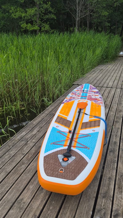 The image shows a paddleboard resting on a wooden pier next to a body of water. The paddleboard is colorful with orange, white, blue, and brown accents, and has a paddle attached. Tall green reeds line the edge of the water, and trees form a dense backdrop. The setting appears to be a natural, outdoor environment. While the location is stated to be Rimšas, Latvia, this specific information is not visually verifiable from the image itself. There are no people or animals visible in the image.