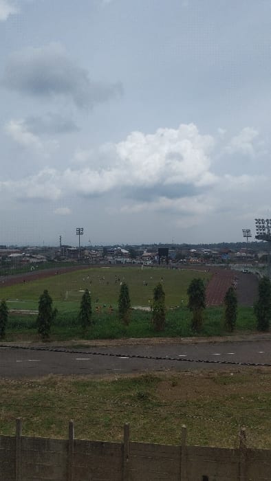 The image shows a view of a soccer field located at latitude: 4.163, longitude: 9.279 in Buea, Cameroon. The field appears to be mostly green with some brown patches and lined with white markings. There are people playing soccer on the field and some trees and bushes surrounding the area. The field is situated between some buildings in the distance, and a few light posts and other structures can be seen surrounding the field. The sky above the field is cloudy, with some white clouds visible against a gray sky. The image appears to have been captured from a higher elevation, looking down at the scene, likely from a nearby building. There are people, a few cars, and structures visible in the distance. There is a large green field in the foreground, with a few trees and bushes surrounding the field.