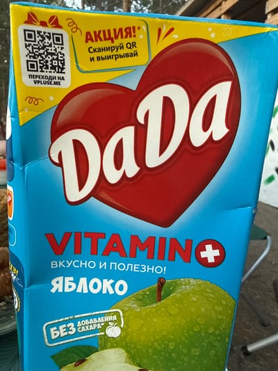 The image displays a juice carton, likely apple-flavored, positioned on its side. The carton features a vibrant design with a bright blue background and a yellow promotional panel on the left. The prominent brand name "DaDa" is emblazoned in white on a large red heart, indicating the product's name.
To the right of the heart, the word "VITAMINO" is vertically oriented, accompanied by a red circle containing a white cross, suggesting the product is vitamin-fortified. Below "VITAMINO," the text "ВКУСНО И ПОЛЕЗНО!" translates to "Tasty and Healthy!". The product's flavor is identified as "ЯБЛОКО" (Apple), illustrated by a fresh green apple adorned with water droplets. A white oval label below the apple explicitly states "БЕЗ ДОБАВЛЕНИЯ САХАРА" (Without added sugar).
The yellow section on the left advertises a "АКЦИЯ!*" (Promotion!). It instructs consumers to "Сканируй QR и выигрывай" (Scan QR and win). A clear QR code is visible, accompanied by the text "ПЕРЕХОДИ НА VPLUSE.ME" (Go to VPLUSE.ME), indicating the promotion's online destination. Decorative stars, swirls, and confetti-like elements adorn this promotional area.
The background is out of focus, suggesting a bright, possibly outdoor or well-lit indoor setting, with blurry hints of wooden structures or foliage. No people are visible. The time of day appears to be daytime, given the bright illumination. The image is contextualized in Zerendi, Kazakhstan. Partially visible text "АХА СЫЙЛЫКТ" (likely "Aha gift" in Kazakh) can be seen at the bottom right.
