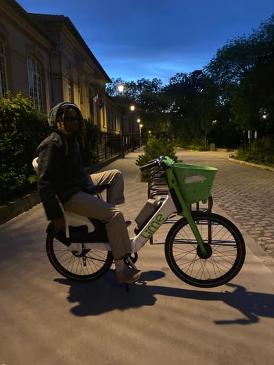 The image shows a person sitting on a Lime bike on a sidewalk in Le Pré-Saint-Gervais, France. The person is wearing a hooded sweatshirt and light-colored pants. The bike is a green and white Lime electric bike. The building is visible in the background, next to the person. The French flag is displayed on the building. The time of day appears to be evening, with a dark blue sky in the background. The pavement is made of cobblestones. The scene is lit by street lights.