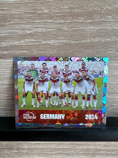 This is a close-up, vertically oriented image of a Panini soccer sticker featuring the Germany national team from the 2014 FIFA World Cup. The sticker has a holographic, diamond-patterned border. The main subject is a team photo of the German players, with eleven players visible, most of whom are in white jerseys with black and red trim, and black shorts. They are posing on a green soccer field with a crowd visible in the background. Some players are standing in two rows, while others are kneeling or sitting. One player, seemingly a goalkeeper, is wearing a green jersey.
Notable details include the year "2014" printed prominently on the right side of the sticker in large white font against a dark red background. Below the year, the words "GERMANY" are vertically oriented in white letters. Further down on the red background, there is a circular logo with the text "FIFA MUSEUM" and a stylized globe. Some of the players have numbers visible on their jerseys, such as "23", "16", "4", "13", and "8". The overall emotion conveyed is one of pride and accomplishment, typical of a team celebrating a victory or a significant tournament. The setting appears to be a professional soccer stadium. The background suggests a sunny day, as indicated by the bright lighting and the presence of a crowd. There is no discernible time of day or weather beyond what is typical for a soccer match. The image is sharp and clear, highlighting the details of the sticker and the players. The sticker itself is in excellent condition, with no visible damage or wear.