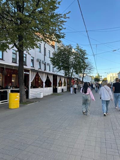 The image depicts a street scene in the city of Feklino, Russia. A pedestrian walkway paved with gray rectangular bricks runs through the center of the frame, lined by buildings and trees. On the left side of the walkway is a restaurant with an outdoor seating area covered by white curtains and string lights. Several people are seated at tables inside and outside the restaurant. A large yellow cylindrical trash bin is positioned next to a tree in front of the restaurant. A group of people, including women and men, are walking away from the camera on the pedestrian walkway. Some of the individuals are dressed in casual clothing, such as jeans and t-shirts. In the background, buildings with white facades and windows are visible, as well as trees and a clear blue sky. Overhead, electrical wires are strung across the sky. The overall impression is of a bustling urban environment with commercial activity and people enjoying a pleasant day.