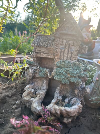 The image depicts a miniature fairy house ornament situated outdoors, likely in a garden setting, with cracked dark soil in the foreground. The fairy house is designed to resemble a rustic dwelling constructed from natural materials, with a textured, wood-like exterior and a roof made to look like thatched straw. Green, moss-like embellishments are visible on the sides of the house, simulating overgrown foliage. Sunlight streams in from the upper right, casting a warm glow and creating lens flare effects. In the background, out-of-focus greenery, including tree branches with long, slender leaves, and a few purple flowers are visible. To the right, partially obscured by the fairy house, a garden gnome statue and other decorative elements can be discerned, suggesting a whimsical or decorative garden arrangement. The scene is set in Velyka Berezovytsia, Ukraine.