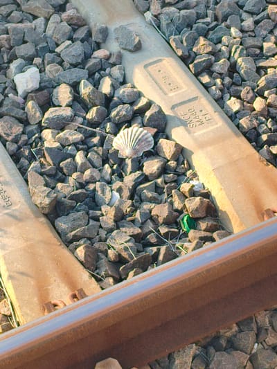 A single scallop shell rests on a bed of ballast between two concrete railway sleepers. The sleepers are marked with text, one reading "SNCF N440 PI" and the other partially obscured with "EGEBA". The foreground features a section of a rusty railway track. The scene appears to be outdoors, with sunlight casting shadows on the rocks and sleepers. The location is identified as Villeneuve-Loubet, France.