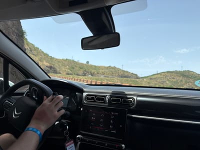 A first-person perspective inside a car shows a person's hands on the steering wheel and interacting with the car's infotainment system. The setting appears to be a sunny day with a clear blue sky, suggesting good weather. Outside the car, a hilly, arid landscape with sparse vegetation is visible through the windshield and side windows, with a road winding through it. The image is taken from the driver's seat, focusing on the dashboard, steering wheel, and the driver's hands.
The driver's left hand is on the steering wheel, which has the Citroën logo. The right hand is reaching towards the center console, where a touchscreen display shows climate control settings, including the temperature set to 21 degrees Celsius and a humidity reading of 29%. The time displayed on the screen is 15:29. A lanyard with a tag that reads "canarias.com" and "Citroën C3" is hanging from the ignition. The driver is also wearing a blue wristband with white polka dots. The overall impression is a casual drive on a clear day, likely in a warm climate given the visible landscape and the climate control settings. The scene is devoid of any strong emotional cues, appearing to be a straightforward depiction of a driving experience.
