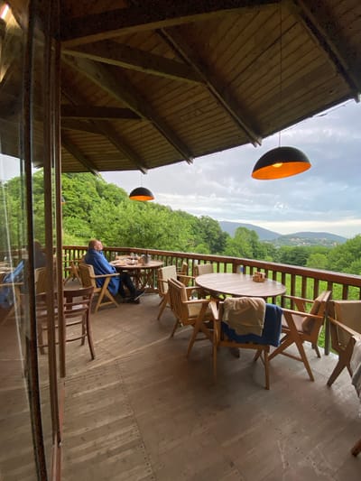 The image shows an outdoor deck with wooden furniture, including chairs and tables. A person is sitting in one of the chairs, wrapped in a blue blanket. The deck overlooks a green, mountainous landscape under a cloudy sky. There are two hanging light fixtures visible under a wooden roof. The scene is viewed through a window. The city of Прогресс, Russia is not evident in the image.