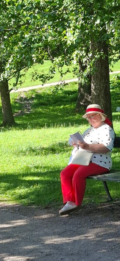A person is sitting on a park bench with a book in their hands, seemingly reading. The individual is wearing a white shirt with black polka dots, red pants, and a straw hat with a red band. They are also wearing glasses. The scene is outdoors, with lush green grass and trees, suggesting a sunny day. The sunlight creates dappled shadows on the grass. The person appears relaxed, engrossed in their reading. The background shows a paved pathway winding through the park, with more trees and greenery. The overall impression is one of a peaceful afternoon spent enjoying a book in nature. The image is oriented vertically, with the person positioned on the right side of the frame, leaning against the bench.