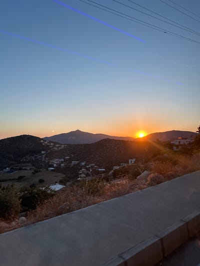 The image captures a beautiful sunset over a mountainous landscape, likely in Kaş, Turkey. The sun, a bright orange and yellow orb, is prominently featured as it dips behind a distant mountain range, casting a warm, glowing light across the horizon.

The sky transitions from a soft, clear blue at the top to vibrant orange and yellow hues near the setting sun. Several faint blue streaks, likely lens flares or light artifacts, are visible diagonally across the blue portion of the sky, particularly in the upper left. Utility lines or cables are also visible stretching across the upper left corner against the clear sky.

The midground is dominated by rolling hills and mountains covered in sparse, Mediterranean-type vegetation, appearing somewhat arid. Scattered across these hillsides are numerous white and light-colored buildings, suggesting a small village or settlement nestled into the terrain.

In the immediate foreground on the right side, a dark, textured concrete or stone wall or railing is visible, indicating the photo was taken from an elevated position, possibly a terrace or balcony. The overall impression is one of a peaceful and serene evening scene. There is no visible text in the image.
