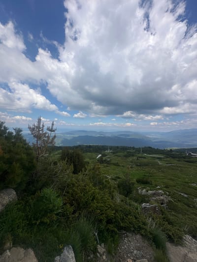 The image depicts a scenic mountain landscape under a partly cloudy sky. In the foreground, there are rocky outcroppings and dense green vegetation, including pine trees and low-lying shrubs. A dry, dead tree stands prominently among the living greenery.
Beyond the immediate foreground, the terrain slopes downwards, revealing a vast expanse of rolling hills and valleys stretching into the distance. The landscape is dotted with trees and patches of green grass, indicative of a mountainous region. In the mid-ground and background, several human-made structures are visible, including what appear to be ski lift towers or telecommunication masts, and a building on the right. The distant horizon shows layers of hazy blue mountains. The sky is a vibrant blue, interspersed with large, fluffy white clouds. The overall impression is one of a remote, natural setting, possibly a mountain resort area.
The exact location in the city of Unknown, Bulgaria, is not discernible from the image. However, the terrain suggests a high-altitude environment.