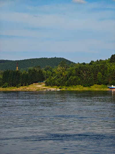The image shows a wide, calm river with ripples on its surface, reflecting the blue sky and scattered clouds. The riverbank is lined with lush green trees, forming a dense forest that stretches up to a rolling hill. On the left side of the riverbank, a tall, narrow industrial chimney with a yellow and brown striped pattern stands out against the green backdrop. There are several small boats pulled up on the sandy shore near the trees. Further to the right, a larger blue and white boat is partially visible on the shore, possibly undergoing maintenance or repairs. The scene appears to be set in Predivinsk, Russia, a city located on the Yenisei River.
