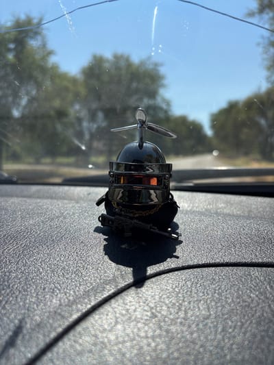 The image shows a car's dashboard with a unique air freshener. The air freshener is a miniature replica of a helicopter, complete with a propeller. It's made of shiny metal and hangs from a small chain, attached to what appears to be a black fabric pouch. The helicopter's body is a dark, reflective sphere with silver detailing. The propeller is also silver and appears to be made of metal.

The air freshener is positioned on the textured surface of the dashboard, casting a sharp shadow. Behind the air freshener, the reflection in the car's windshield shows a blurred landscape of trees and a clear blue sky, suggesting it's daytime. The sun is bright, creating highlights on the windshield and the air freshener. The overall scene is one of a sunny day, likely in a rural or suburban setting, as indicated by the greenery visible through the windshield. There is no text visible in the image. The focus is on the novelty of the air freshener.