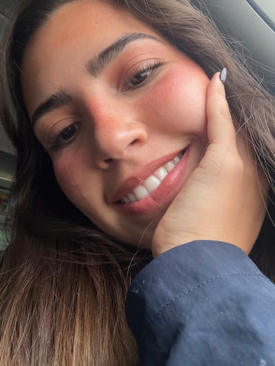 A close-up, rotated portrait shot depicts a person with light skin, dark eyebrows, and brown hair. The individual is smiling, revealing straight, white teeth. Their hand is positioned against their cheek, with a visible gray nail polish. A dark blue sleeve is also present in the frame. No information in the image allows to deduce a location.