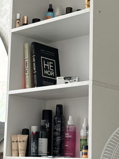 The image depicts a white, multi-tiered shelf unit filled with various items. The top shelf holds several cosmetic and personal care products, including small bottles and jars, as well as a black container. A blue spray bottle with a gold-colored label is also visible. The second shelf down features two books standing upright, one with a black cover and white text, and another with a lighter spine. Next to the books is a small silver digital camera with a lens. The third shelf is densely packed with an assortment of cosmetic products, including tubes, bottles, and spray cans, in various sizes and colors, some with visible labels in Cyrillic. A small fan is partially visible on the right side of the image, suggesting it is in a room. There are no people or any indications of the city of Червишево, Russia.