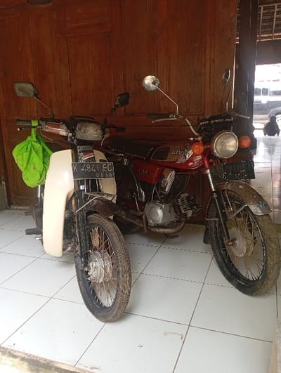 Two motorcycles are parked side-by-side on a white tiled floor within a covered area, presumably a garage or porch, in Kajar, Indonesia. A wooden paneled wall forms the immediate background.

The motorcycle on the left is an underbone type with a cream-colored front fairing and a black front fender. Its front license plate, "K 4841 EC," with an "01 19" designation below, is visible. A bright green fabric bag hangs from its left handlebar. It features spoked wheels with black rims.

The motorcycle on the right is a red Yamaha, appearing to be an older model, distinguished by its prominent round headlight and "YAMAHA" branding on the fuel tank. It has chrome-finished engine components and handlebars. Its front fender is dark and appears soiled. This motorcycle also has spoked wheels with black rims.

Beyond the wooden wall to the far right, an outdoor area is visible, including a paved surface, a dark vehicle, and a human figure in a crouched posture. Overhead, structural elements consistent with a roof are observable.