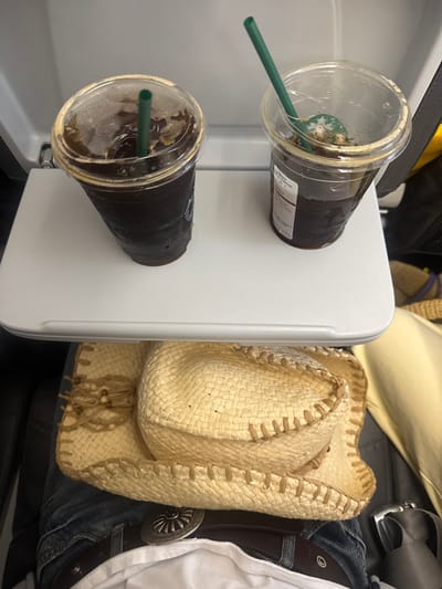 The image depicts an airplane cabin interior. Two clear plastic cups, each containing a dark, iced beverage with a green straw, rest on an extended light-gray tray table. The left cup is mostly full and exhibits condensation. The right cup is approximately half-full, also with condensation, and contains a green stirrer alongside its straw, with a white label visible on its side. Below the tray table, a person's lap is visible, clad in blue jeans, a dark brown belt with a prominent circular silver buckle featuring radial designs, and a white garment. A light-colored straw cowboy hat with a beaded band and textured stitching lies on the lap. The background shows dark seat upholstery and indistinct cabin elements. This scene is presented as occurring within the city of San Miguel Jaltocan, Mexico.