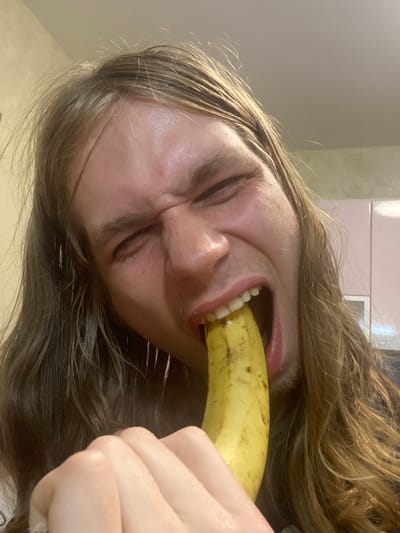 The image is a close-up, self-taken photograph of a person with long, brown hair, biting into a banana. The person's face is contorted in an expression of effort or intensity, with their eyes squinted and mouth wide open around the banana. Their hand is visible, holding the banana near their mouth. The background appears to be an indoor setting, possibly a kitchen, with what looks like light-colored cabinets and a wall. There is no indication of the location being Lymanka, Ukraine.