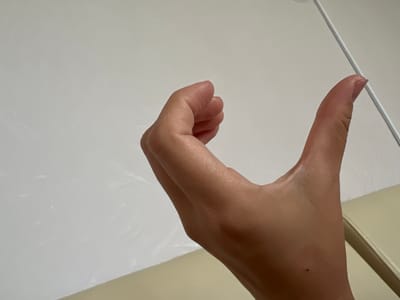 The image shows a close-up of a person's hand, held in a gesture that resembles a letter U or a partial embrace. The hand is positioned against a plain, light-colored background, possibly a wall or a sheet. The skin tone is fair to medium. The thumb is extended upwards, while the fingers are curled inwards, creating a concave shape. A thin, white object, possibly a cable or a ruler, runs diagonally in the upper right portion of the frame. The lower part of the image shows a glimpse of a cream-colored surface, possibly furniture. The lighting is soft and diffused. The location in the city of Үшарал, Kazakhstan cannot be determined from the visual information provided in the image.
